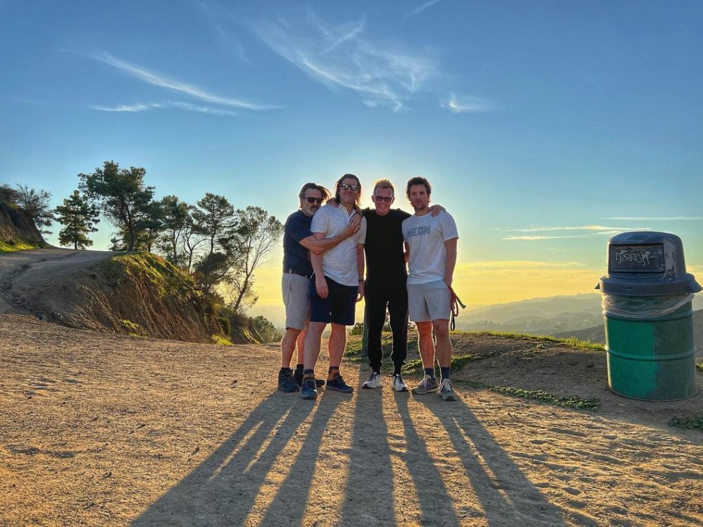 Con O'Neill stands with three friends. He is the person at the very left of the group. He has his arms around an unknown male-presenting person. The next man is Stephen Ward, the person who posted this picture to Instagram. Another unknown male-presenting person stands at the very right. Behind them are rolling hills and a setting sun.