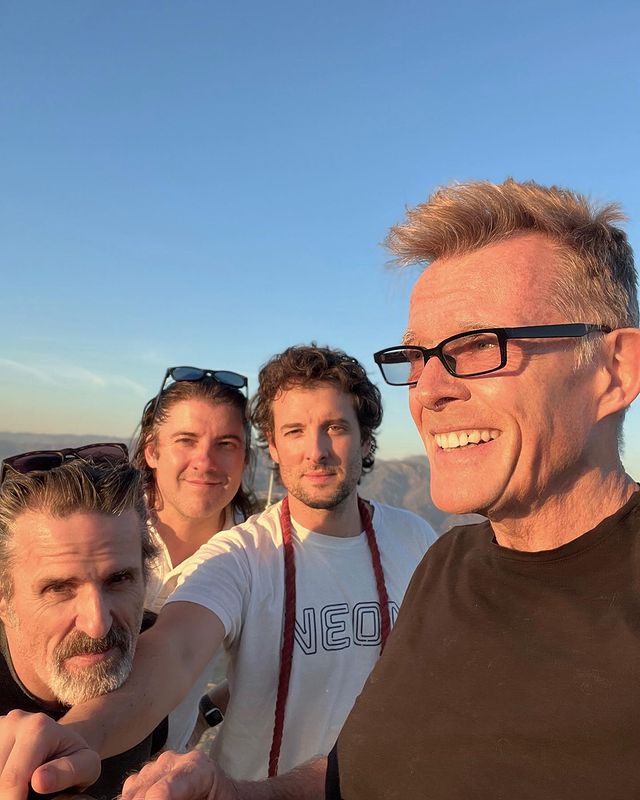 Con O'Neill stands to the left of three other male-presenting people. The very right-hand person is Stephen Ward, the person who posted the Instagram. Stephen is looking towards a setting sun. The others look to the camera. Behind them are rolling hills and a clear blue sky.