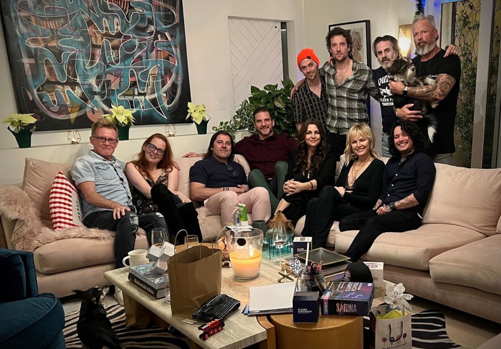 Con O'Neill poses with ten friends, three standing (along with Con) and seven sitting on a pink couch. There are white poinsettia along a ledge behind the sofa. The location tag on this Instagram post said this was taken in Hollywood Hills.