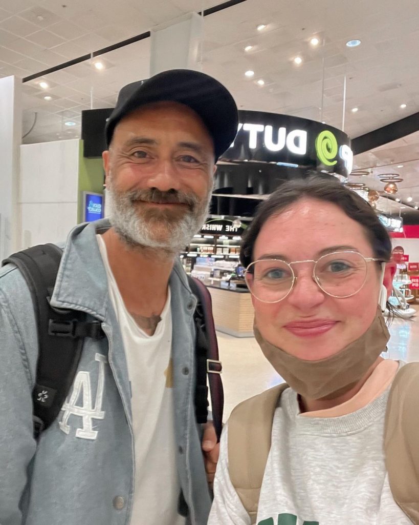 Taika Waititi, wearing a hat and jean jacket, takes a selfie with a female-presenting fan in an airport