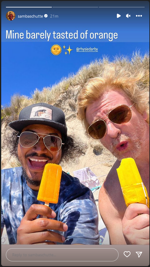 Samba and Rhys sit on a beach, sandy hill behind them, as they eat ice pops.