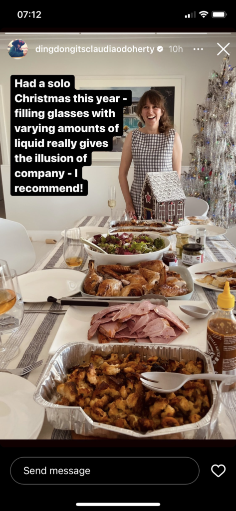 Claudia O'Doherty stands cheerily in front of a large dining table full of traditional Christmas foods like ham, stuffing, and a gingerbread house. Text says "Had a solo Christmas this year - filling glasses with varying amounts of liquid really gives the illusion of company - I recommend!"