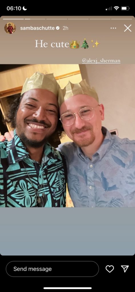 Samba Schutte stands with an arm around OFMD writer Alex Sherman. Both are wearing gold paper crowns from Christmas crackers. Overlay text says: "He cute 👑🎄✨"