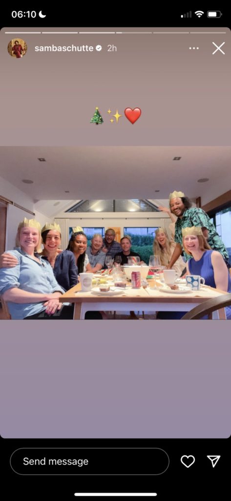 Samba Schutte stands by a large dining table. Either other people are seated around it, including his partner Aria, OFMD creator David Jenkins, and OFMD writer Alex Sherman. Overlay text says: "🎄✨❤"