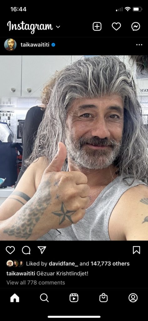 Taika Waititi has his fake Blackbeard wig too far onto his forehead. It looks like he has an 80s pompadour. Taika is giving a thumbs up and has tattoos on his hand (but not on his arms).