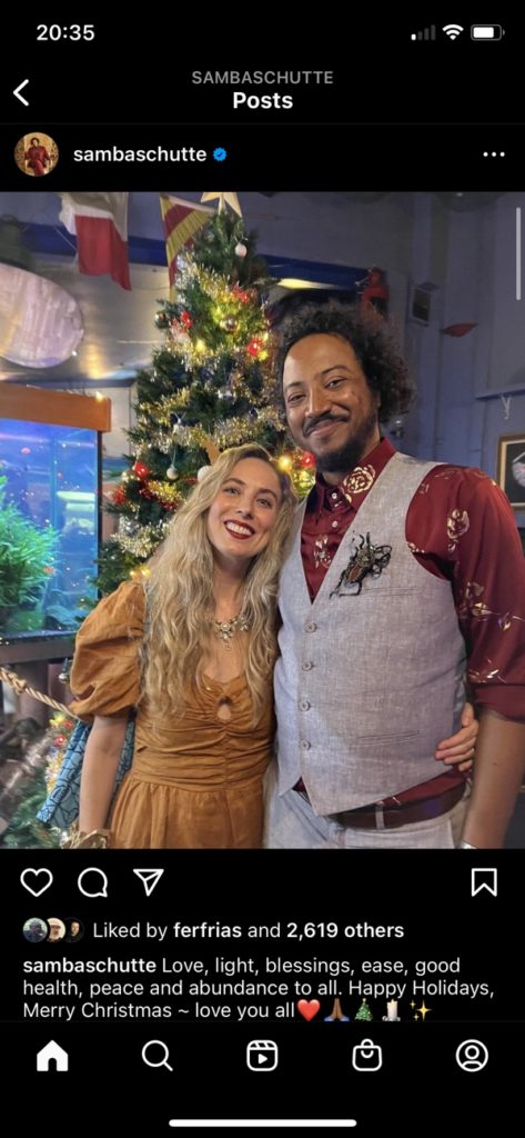 Samba poses with his partner Aria Bruss in front of a Christmas tree. Caption: "Love, light, blessings, ease, good health, peace and abundance to all. Happy Holidays, Merry Christmas ~ love you all❤️🙏🏾🎄🕯✨"