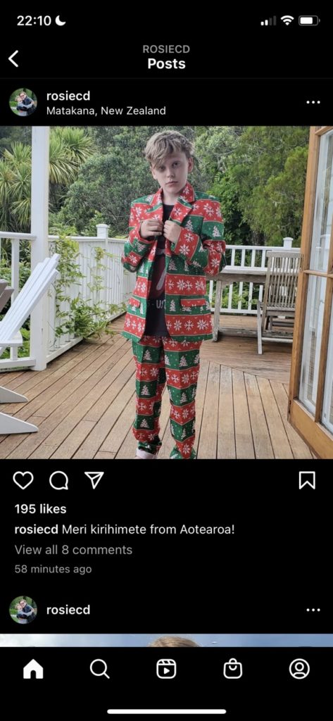 Rhys' son Theo stands on the front porch, wearing a Christmas-themed suit. It has horizontal stripes of alternating red and green with Christmas trees and snowflakes on it. (It's fabulous.) Text caption reads: "Meri kirihimete from Aotearoa!"