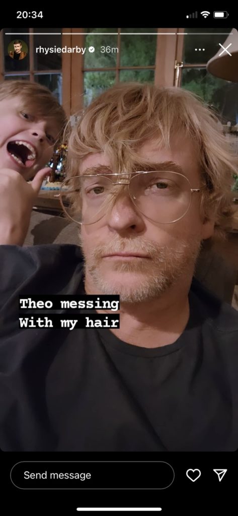 Rhys looks at the camera with resignation as his son Theo smiles goofily behind him, pointing at a lock of Rhys' hair that has been shoved under the bridge of his glasses. Overlay text says: "Theo messing with my hair"