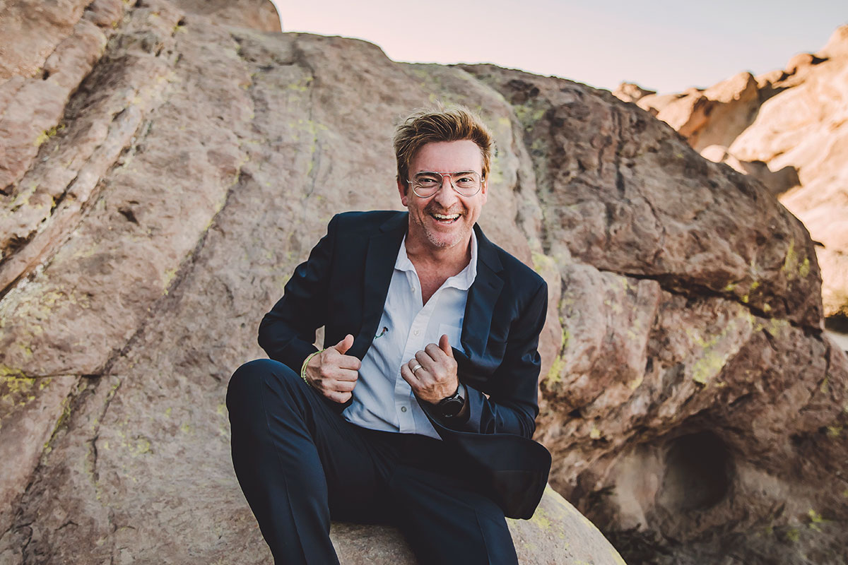 Rhys Darby gives a double thumbs up and a wide smile atop some high desert rocks