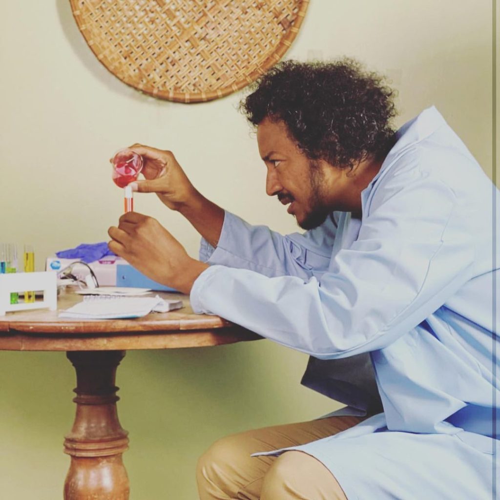 Samba sits, wearing a blue lab coat, at a round wooden table with boxes and papers and test tubes holding colored liquids. He’s pouring something from a beaker into a test tube.