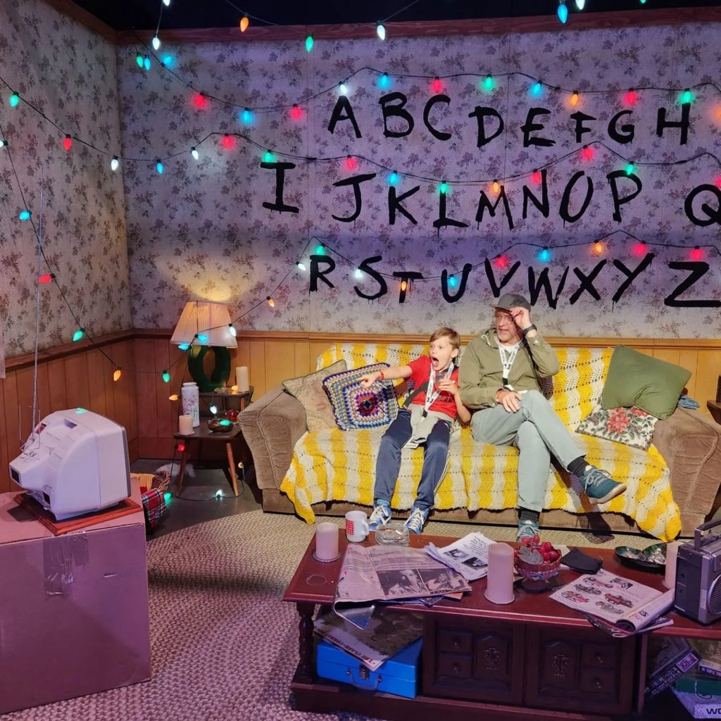 Rhys and son Theo sit on the couch in a replica "Stranger Things" set (complete with alphabet and Christmas lights on the wall). Theo is excitedly pointing to something on a TV across the room.