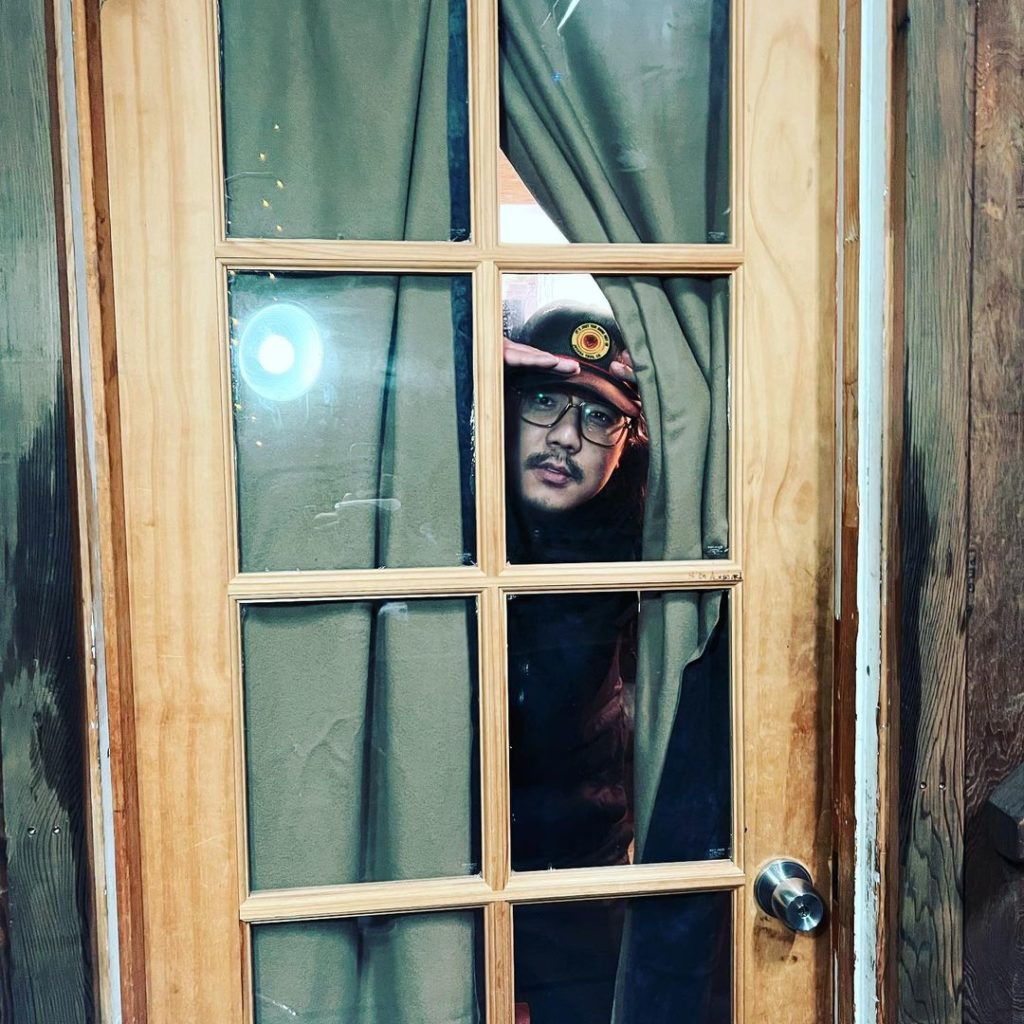 An Asian male-presenting person, wearing a baseball cap, peers at the camera through a pane of glass in a door.