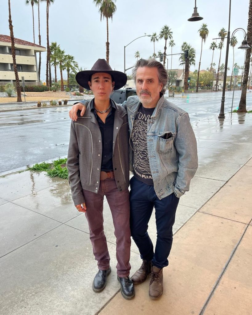 Vico stands next to Con, Con has his arm around Vico’s shoulders. Vico looks stunning in a modern version of a cowboy outfit. Con is wearing a casual jean jacket and jeans.