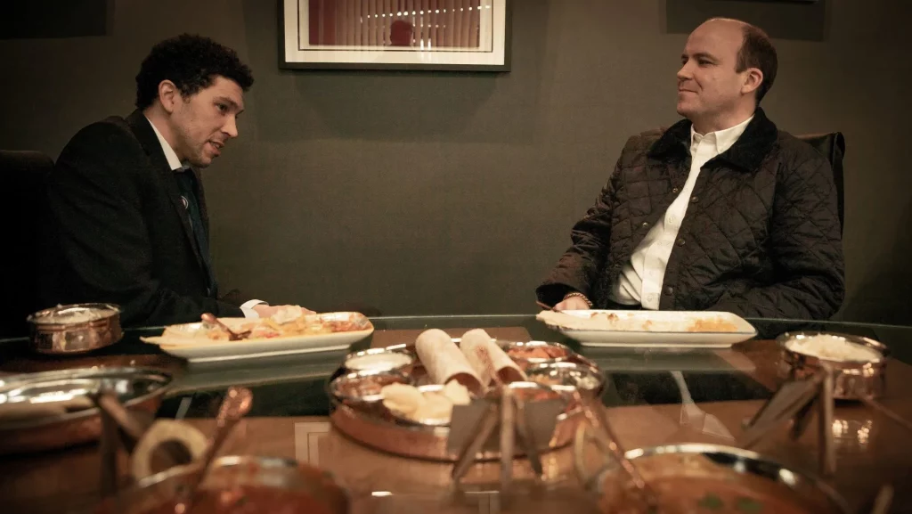 Still from Netflix's "Bank of Dave." Joel Fry and Rory Kinnear's characters sit together at a table eating a meal.