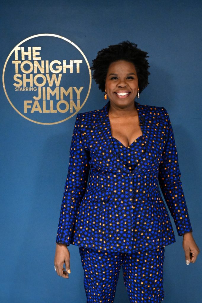 Leslie Jones stands in front of the "Tonight Show Starring Jimmy Fallon" logo on a blue wall. She is wearing a blue dress with orange polka-dots.