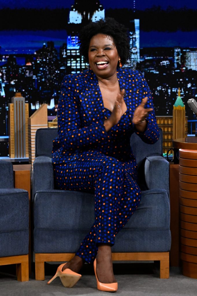 Leslie Jones sits in a guest chair on "Tonight Show Starring Jimmy Fallon". She is clapping and laughing, and wearing a blue dress with orange polka-dots and orange heels.