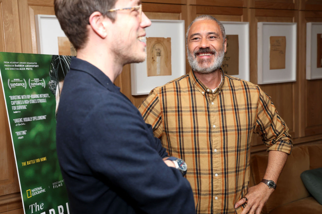 Taika Waititi laughs with Alex Pritz (Director), a white man with short hair and glasses.