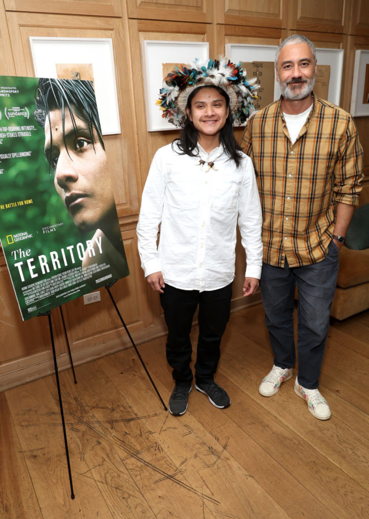 Taika Waititi stands next to Bitaté Uru Eu Wau Wau, who in turn stands next to a movie poster of "The Territory," a film about his people. Bitaté is wearing a white button-up shirt and a traditional flower crown.