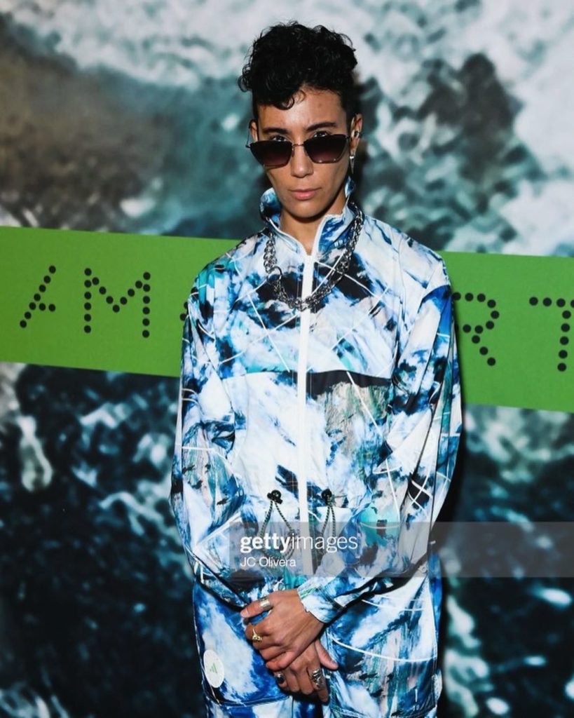 Vico stands in front of the “Stella McCartney” logo for Getty Photos. Wearing a blue textured tracksuit and sunglasses, they strike a power pose, clapping their hands in front of them and looking over their sunglasses at the photographer.