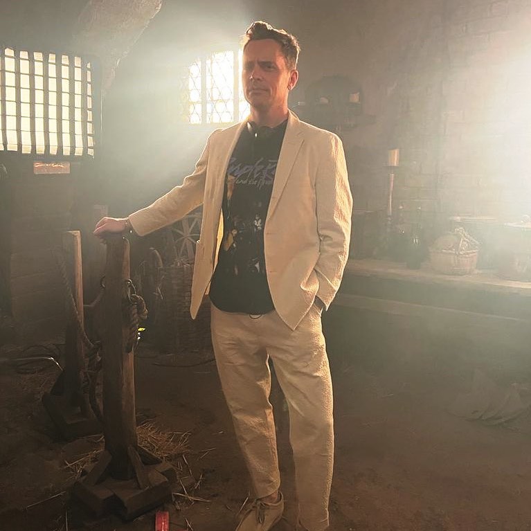 David Jenkins stands in a dark and smoky room, a hand on what looks to be a horse's hitch, a shelf in the background with bottles and pots and candles. David himself is wearing a white suit and black "Purple Rain" t-shirt.