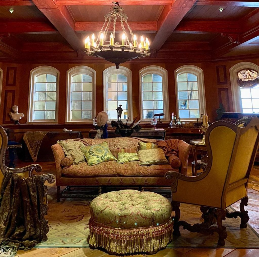 Behind the scenes from S1. Stede's bedroom on the Revenge. There is a couch with two armchairs sitting across, and a round ottoman between. There is a desk behind, and some knick-knacks lining a long shelf running underneath some windows.