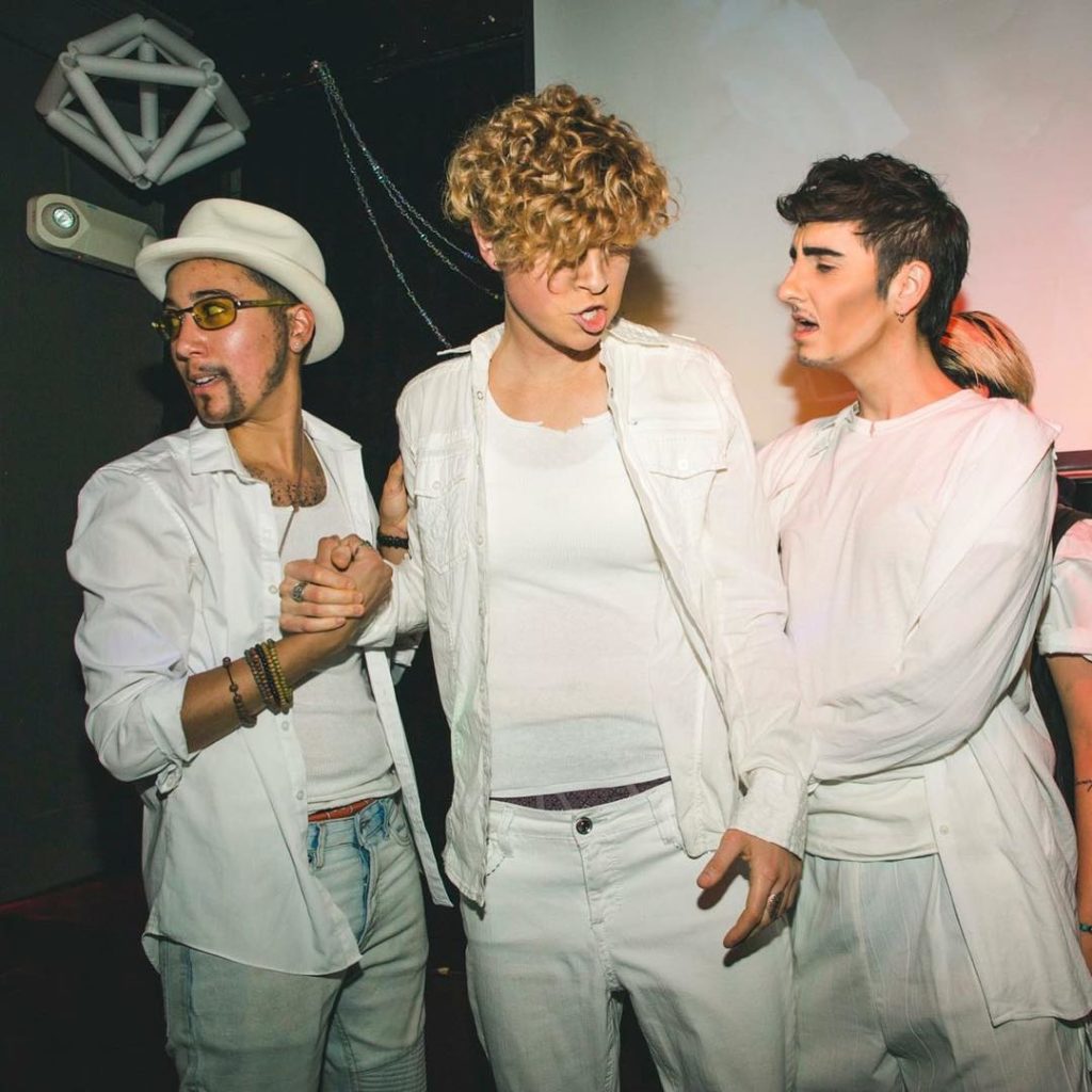 Three drag kings pose as Backstreet Boys members, with their famous all-white outfits. Vico is at the far left, wearing a white hat, yellow-tinted sunglasses, and false goatee; they are looking off-camera while doing a "dap handshake" (I don't know if that's the right phrase) with the middle person.