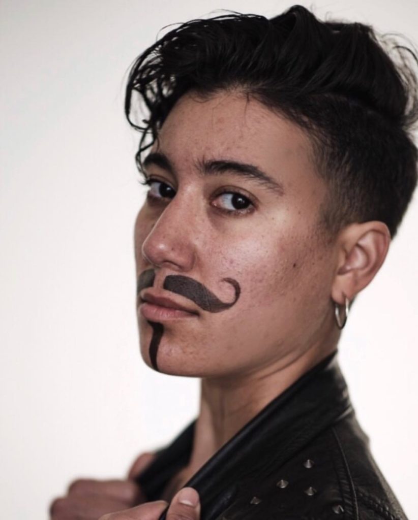 A headshot of Vico. They are wearing a leather jacket and gold hoop earrings, and have a false mustache and goatee on.