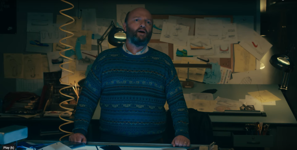 Screenshot from "Air" trailer. Matthew Maher stands there looking like a snack in a sweater. He's asking a question. Nike designs are pinned up on a board behind him.