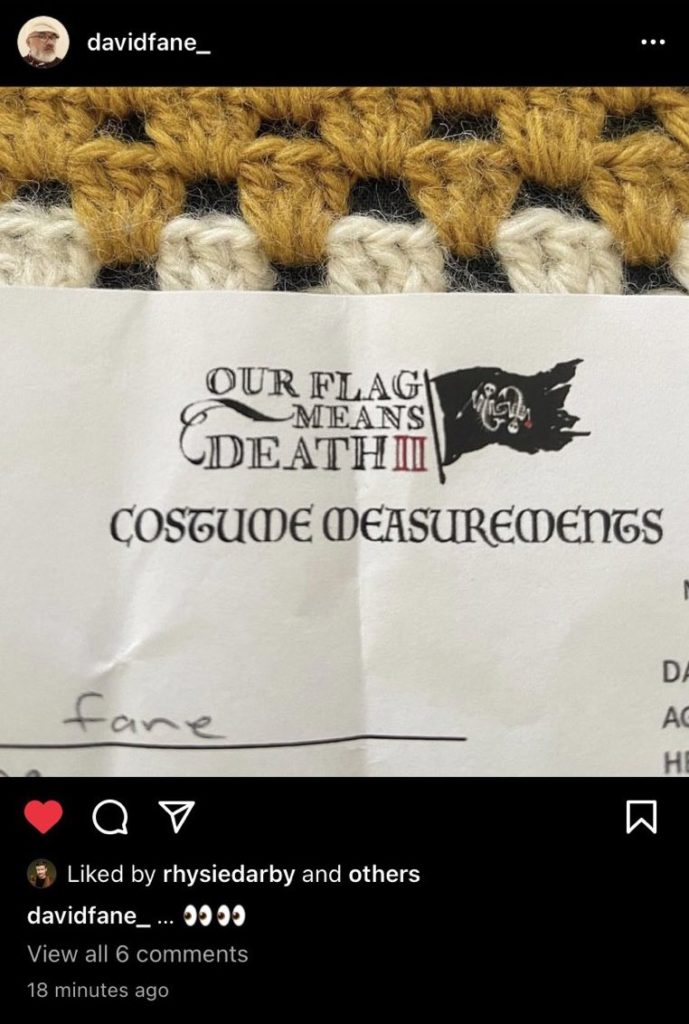 A piece of paper. At the top, there's the words "Our Flag Means Death II" (2 is written with two Is) and a black flag flying next to it. The flag has on it two skeleton mermaids swimming in circles around each other, one holding Blackbeard's spear and the other the heart from Blackbeard's flag. Underneath the title and flag are the words "Costume Measurements." David's caption is: "👀👀". Rhys has liked the photo.