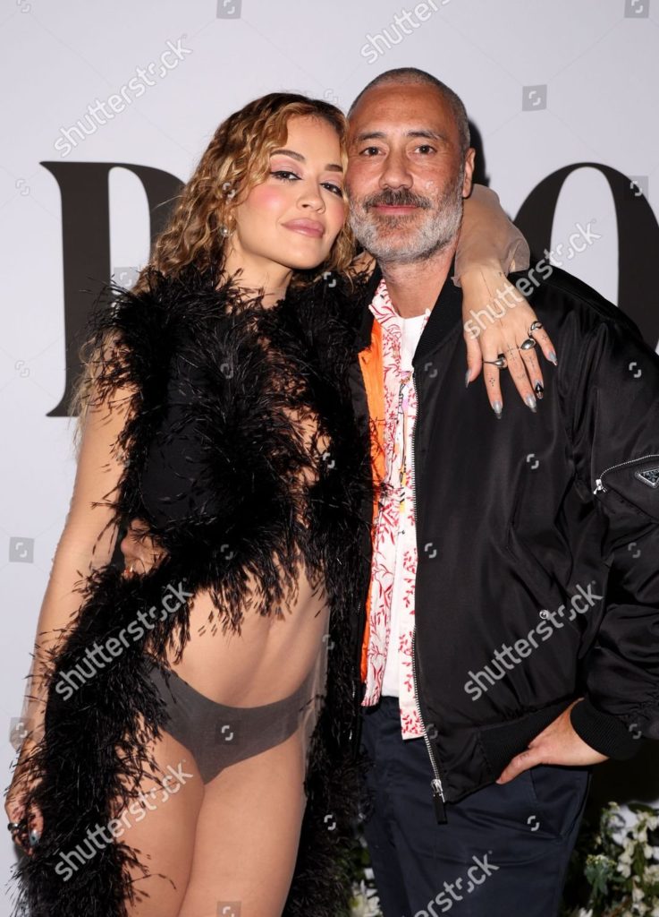 Rita, scantily-clad, drapes an arm around husband Taika, who is wearing a sports jacket and relaxed clothing.