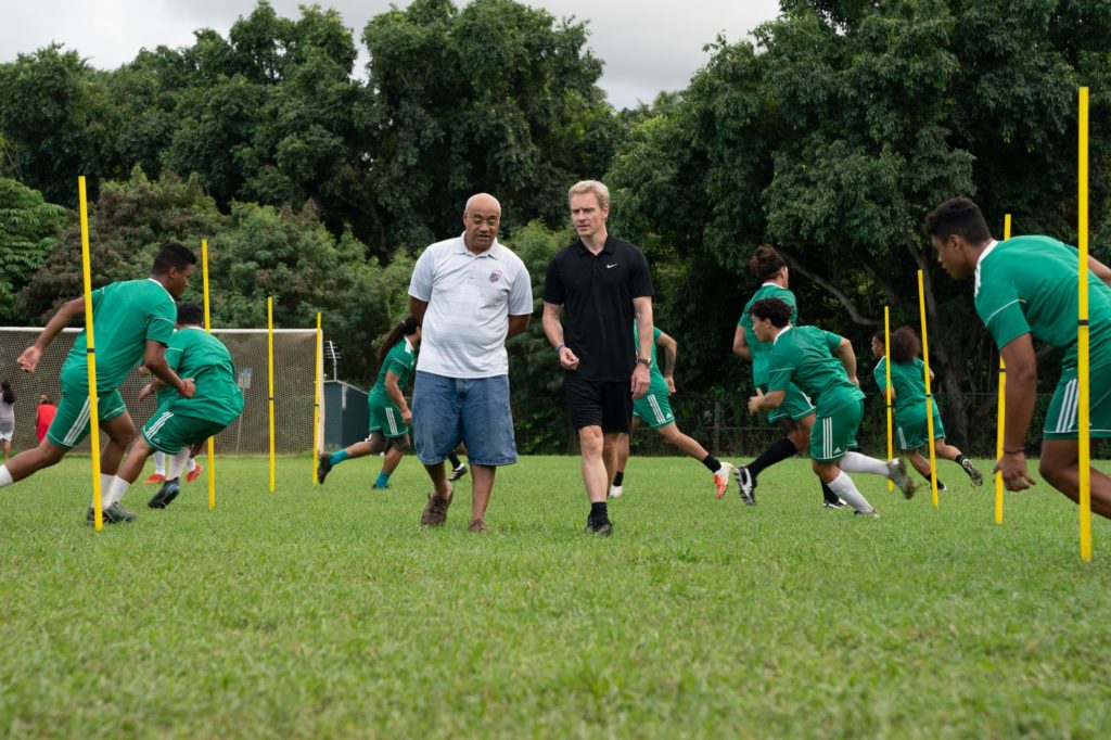 Screenshot from "Next Goal Wins." Michael Fassbender walks next to a presumptive rugby coach, while children play rugby in the background.